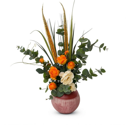 A floral arrangement featuring orange roses, a cream rose, green eucalyptus leaves, and dried golden ornamental grasses in a textured pinkish-red round ceramic vase, set against a plain white background.