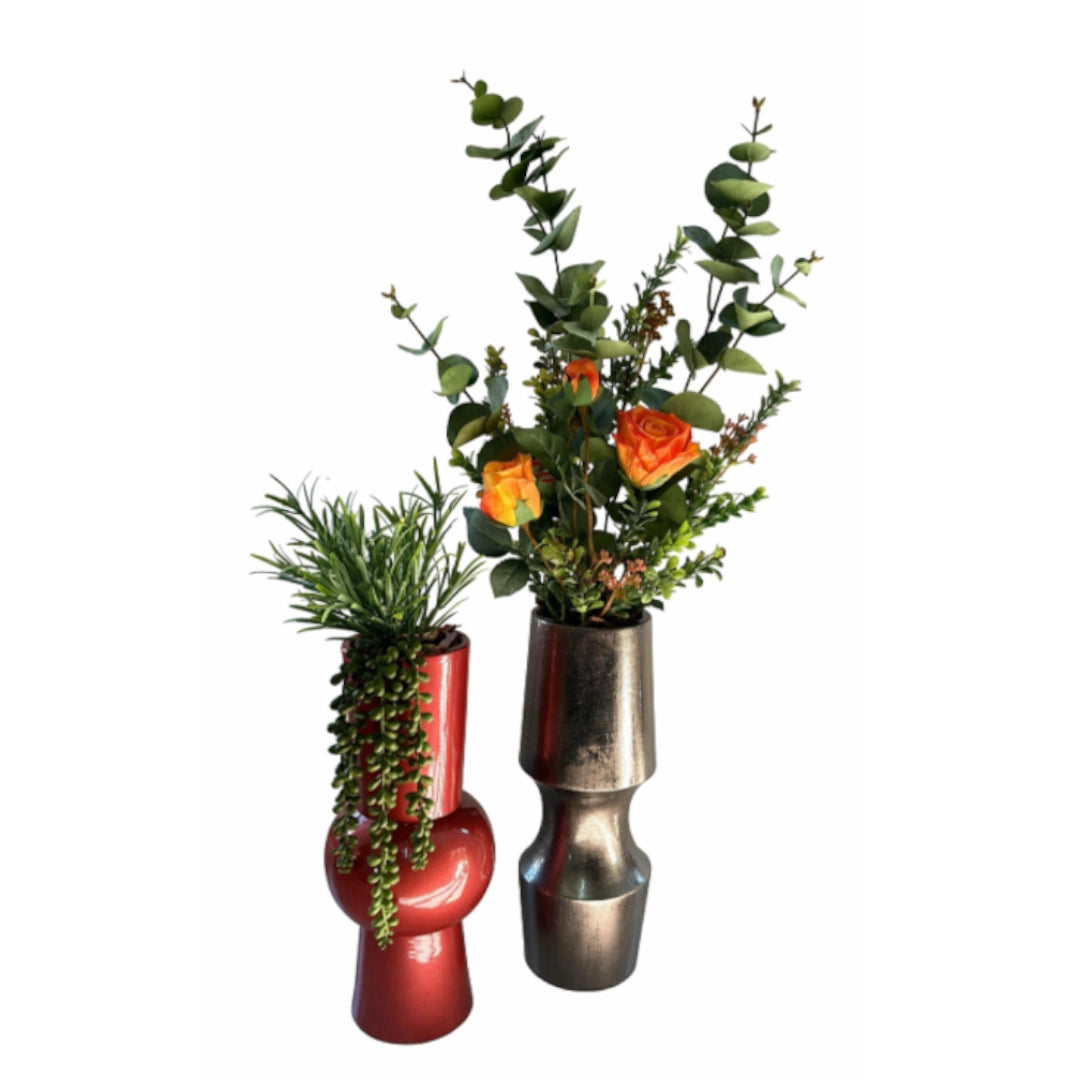 Two decorative ceramic vases with sculptural shapes, one glossy red and one metallic silver, filled with green foliage and orange flowers, set against a plain white background.