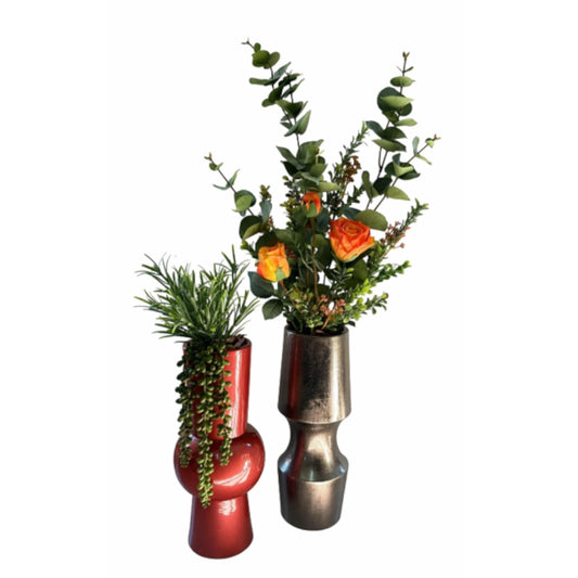 Two decorative ceramic vases with sculptural shapes, one glossy red and one metallic silver, filled with green foliage and orange flowers, set against a plain white background.