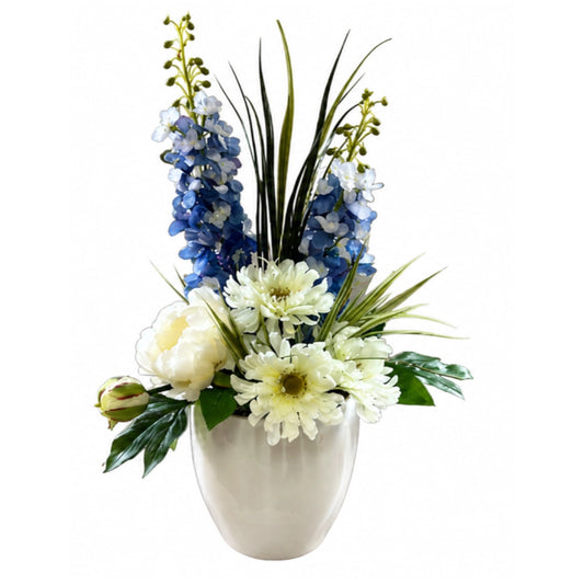An artificial floral arrangement featuring blue delphiniums, white gerbera daisies, white peonies, and green foliage in a glossy white ceramic vase, isolated on a white background.